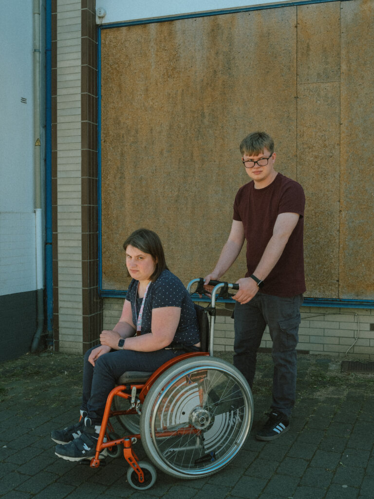 Das Bild zeigt Muriel Sievers in einem Rollstuhl. Daneben steht Nick Becker. Er hält die Griffe des Rollstuhls. Sie sehen mit ernstem Blick in die Kamera.