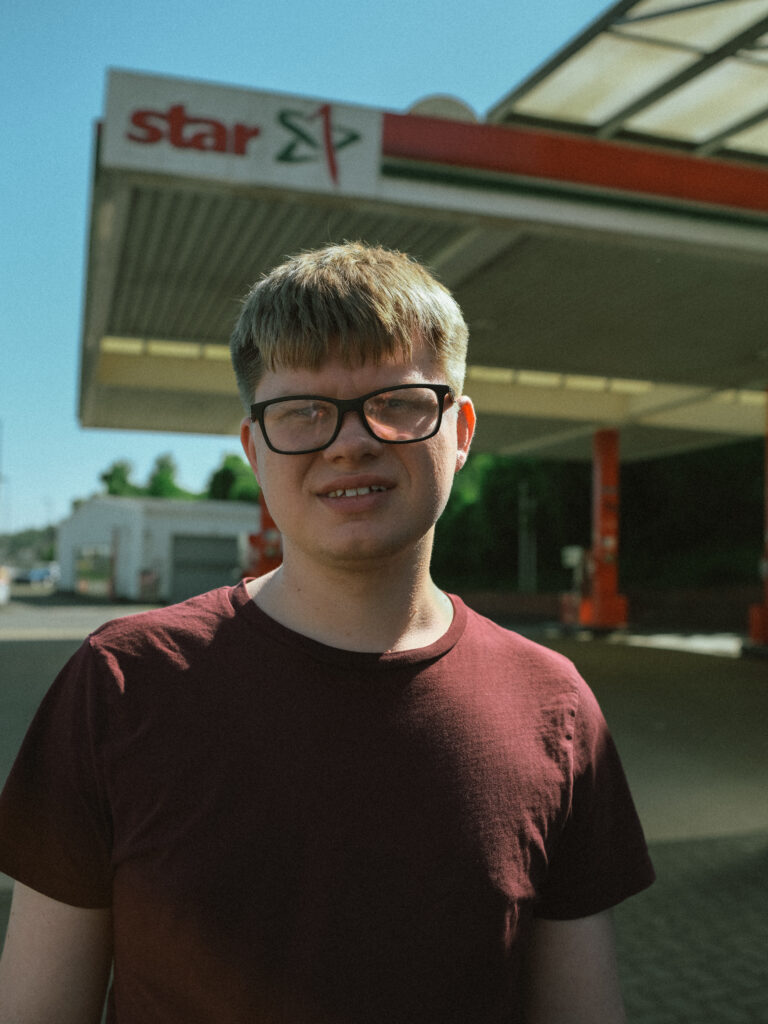 Ein junger Mann schaut mit ernstem Blick in die Kamera. Er trägt eine Brille und kurze blonde Haare. Im Hintergrund sieht man eine Tankstelle.