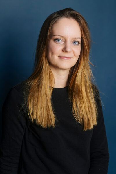 Eine junge, Professionell gekleidete Frau mit Blonden Haaren blickt in die Kamera.