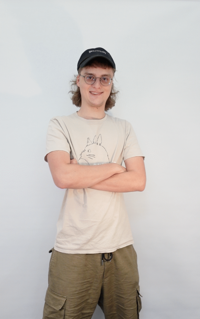 Johannes hat ein weißen T-Shirt und eine schwarze Kappe an. Er hat seine Arme verschränkt und trägt kurze, blonde Haare.