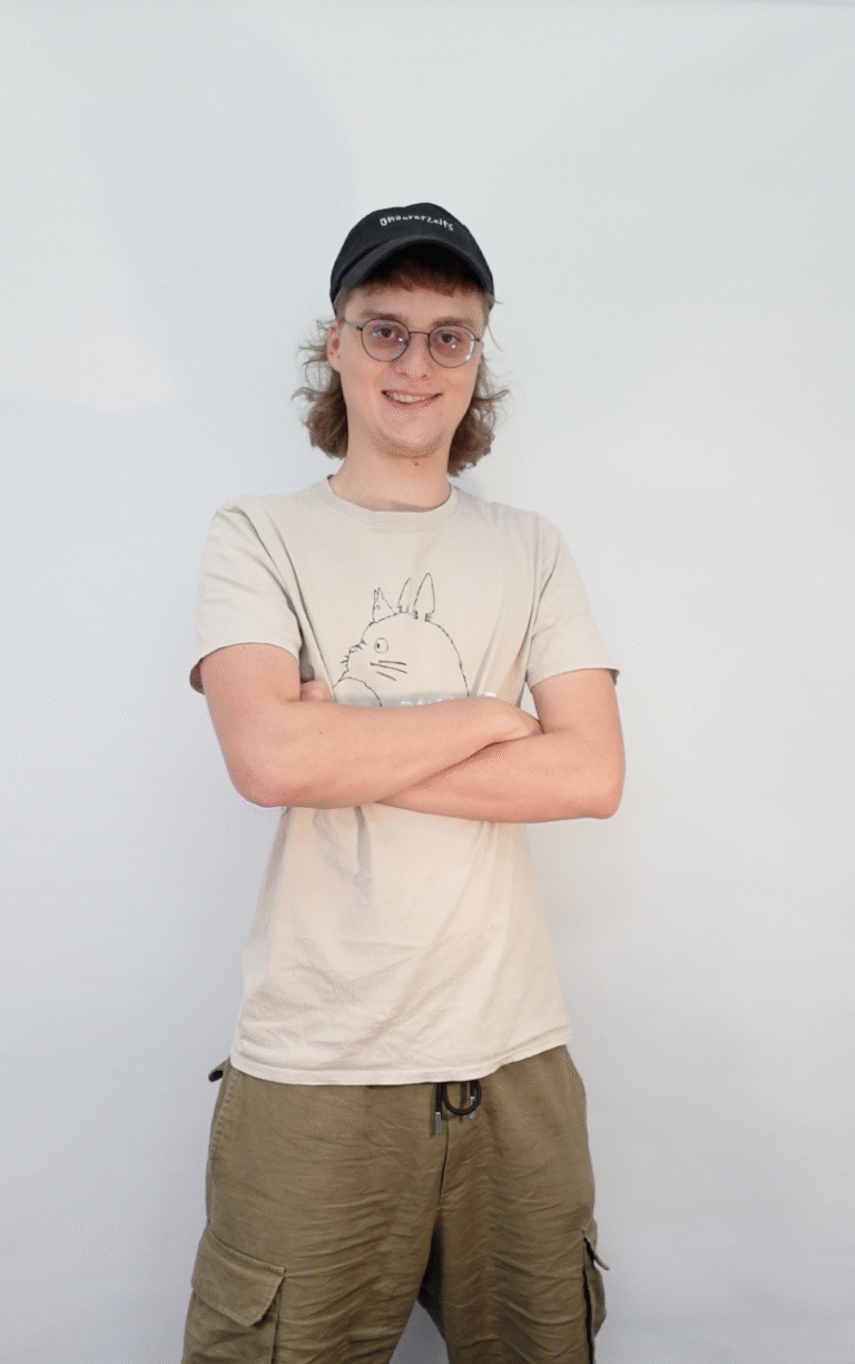 Johannes hat ein weißen T-Shirt und eine schwarze Kappe an. Er hat seine Arme verschränkt und trägt kurze, blonde Haare.