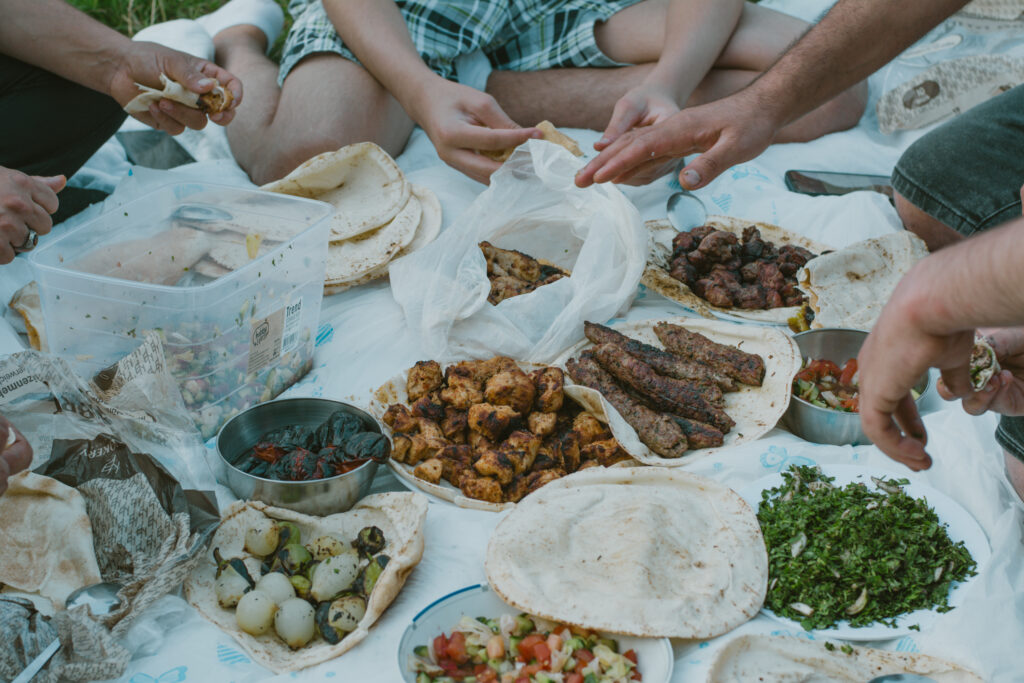 Mehrere Menschen sitzen auf einer Decke und essen zusammen. Auf dem Tuch liegen viele Schüsseln und Teller mit Fleisch, Brot, Salat und Gemüse.