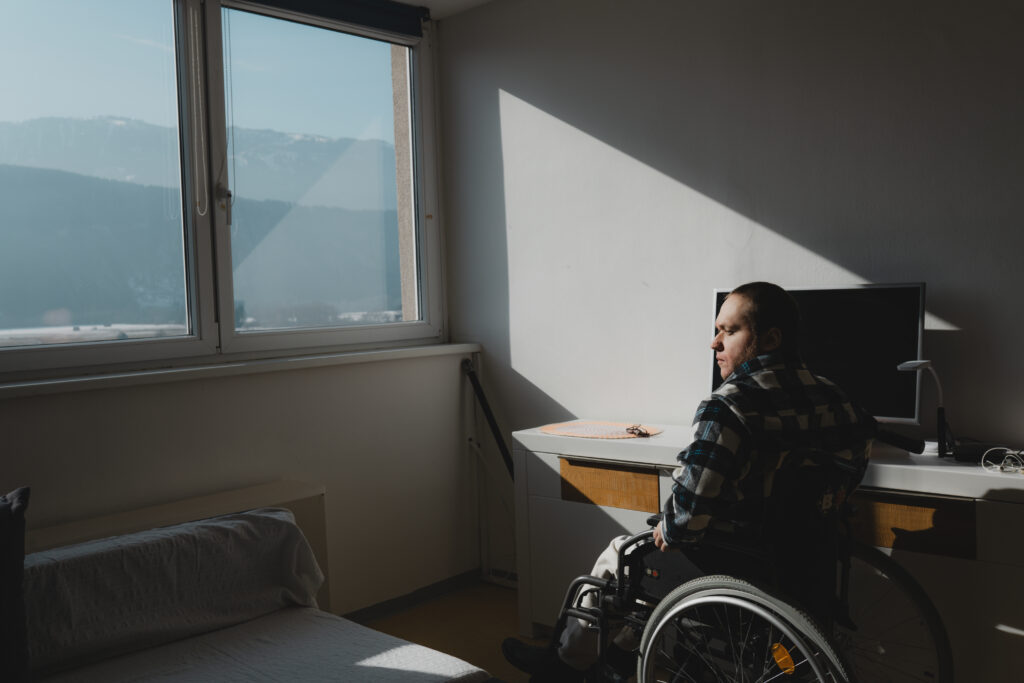 Ein junger Mann sitzt in einem Rollstuhl. Er befindet sich in einer Wohnung. Er schaut aus dem Fenster auf Berge.