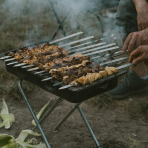 Auf einem Griller liegen mehrere Fleisch-Spieße. An der rechten Ecke sieht man die Hände von mehreren Personen, die nach den Spießen greifen.