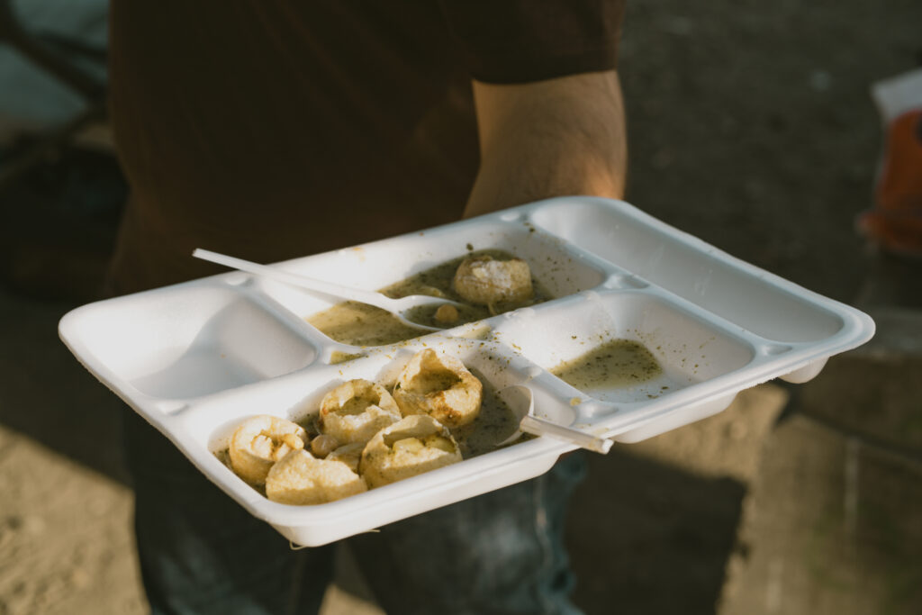 Eine Person hält ein weißes Plastik-Tablett mit Essen. Darin liegen runde Snacks mit Soße und zwei Löffel.