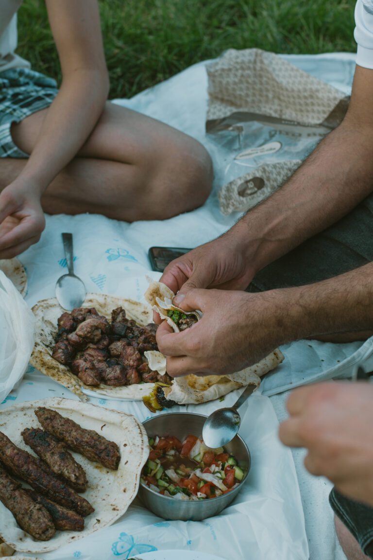 Das Foto zeigt ein Picknick. Auf einer weißen Decke sitzen Personen. In der Hand halten sie Fladenbrot. Auf der Decke ist ein Salat und Lammspieße.