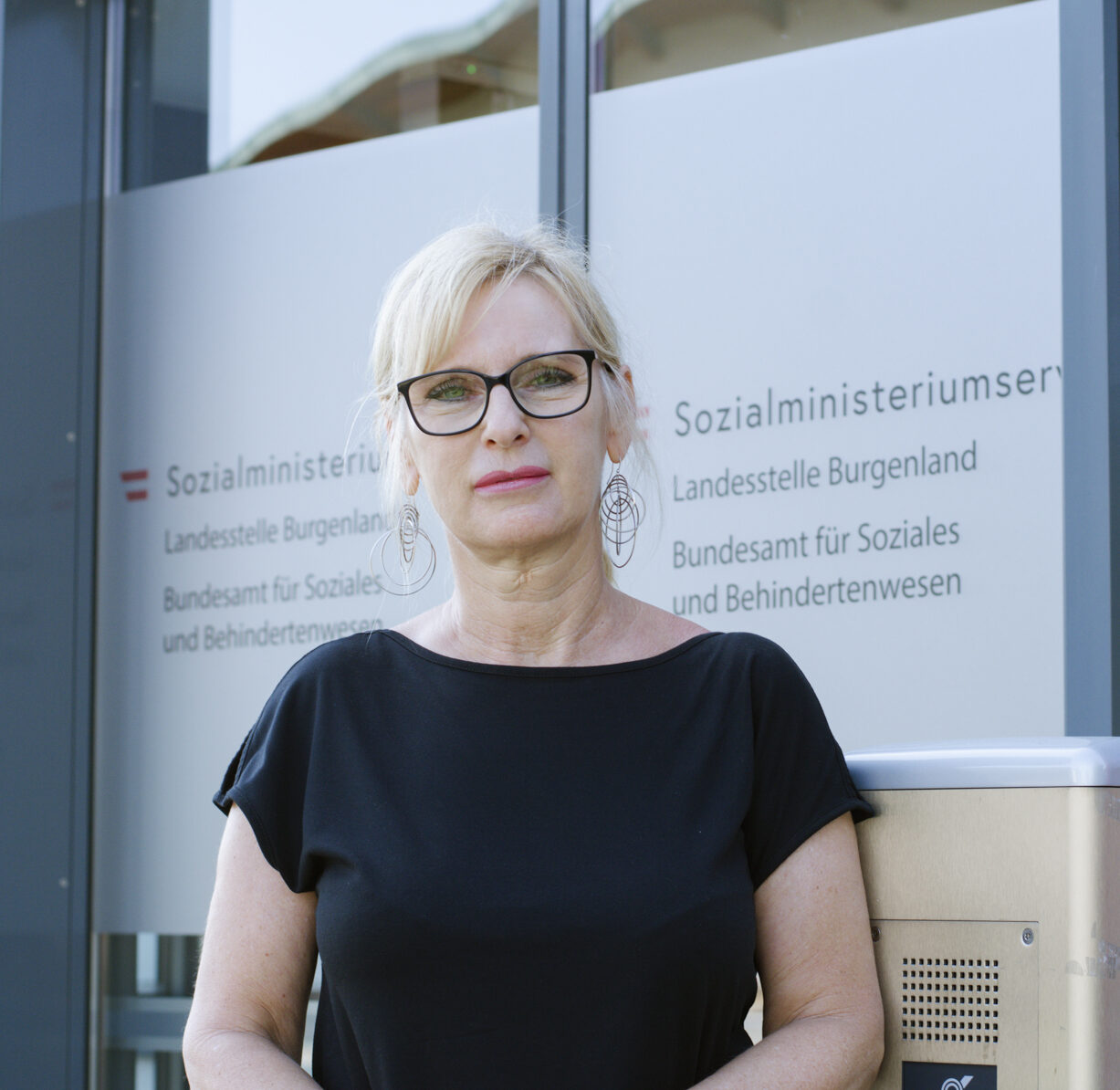 Iris Hübner steht vor dem Sozialministerium und schaut in die Kamera. Sie ist 55 Jahre alt, hat blonde Haare und trägt eine Brille.