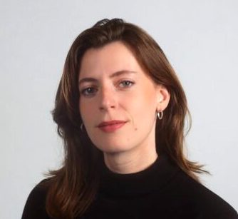 Eine Frau schaut mit ernstem Blick in die Kamera. Sie hat braune Haare und trägt einen schwarzen Pullover.