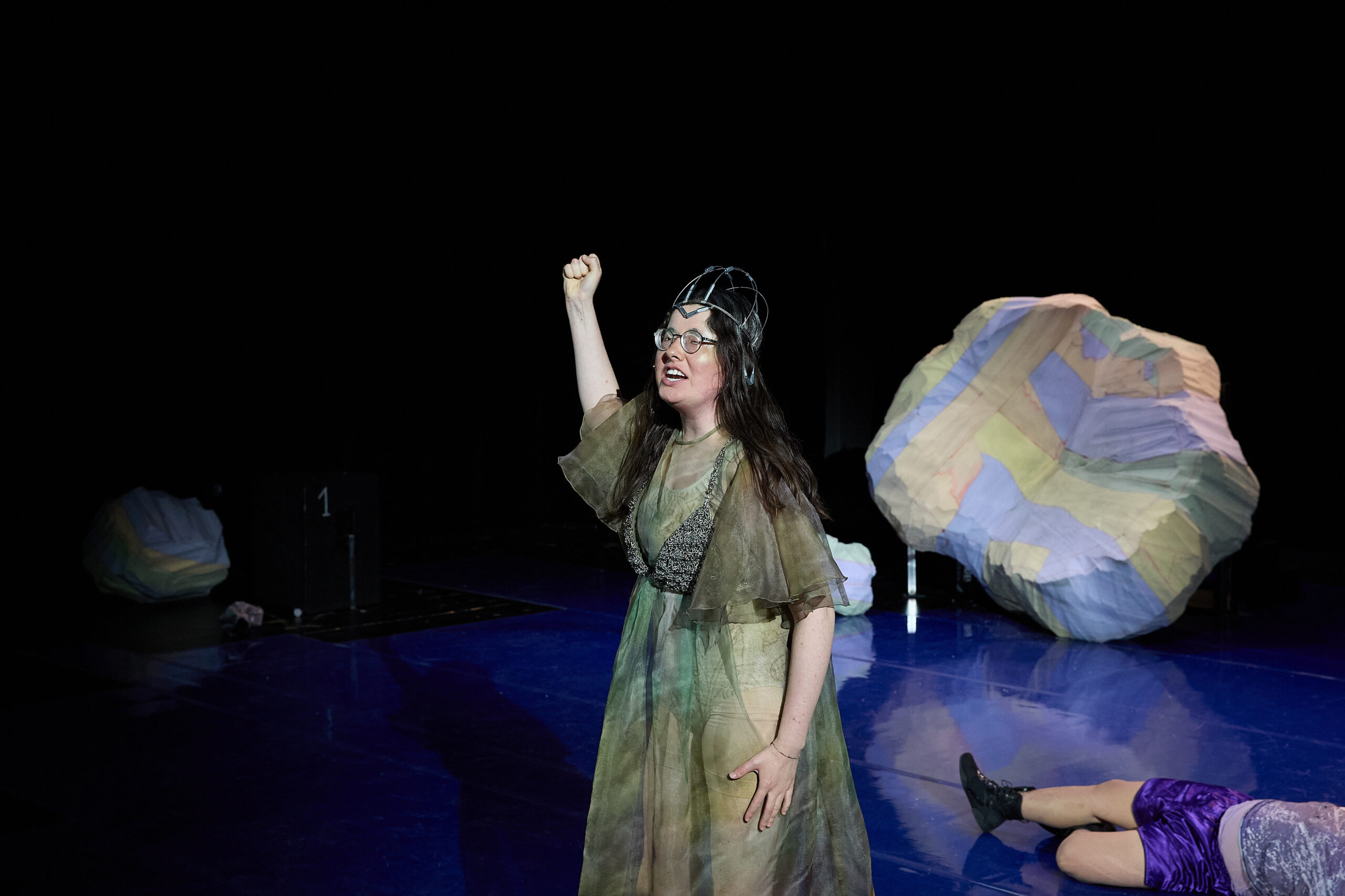 Eine Frau steht auf einer Bühne. Sie hält die rechte Hand zur Faust geballt. Sie trägt ein grünes Kleid. Der Hintergrund ist blau.