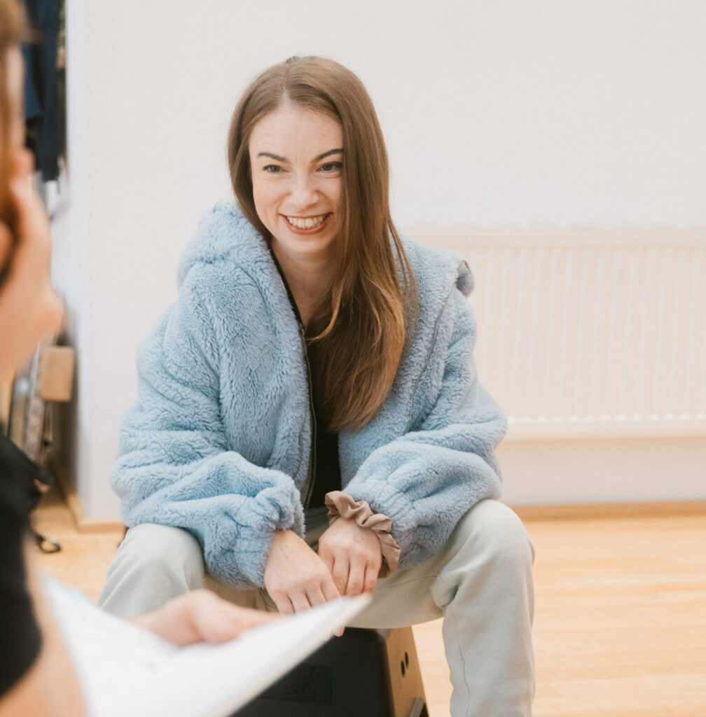 Eine junge Frau lächelt eine andere Person an. Die junge Frau hat lange Haare und trägt eine hellblaue Jacke.