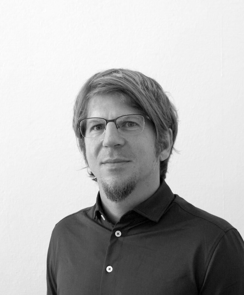 Ein Mann mit kurzen Haaren und Brille schaut ernst in die Kamera. Er trägt ein schwarzes Hemd. Das Bild ist in schwarz weiss.