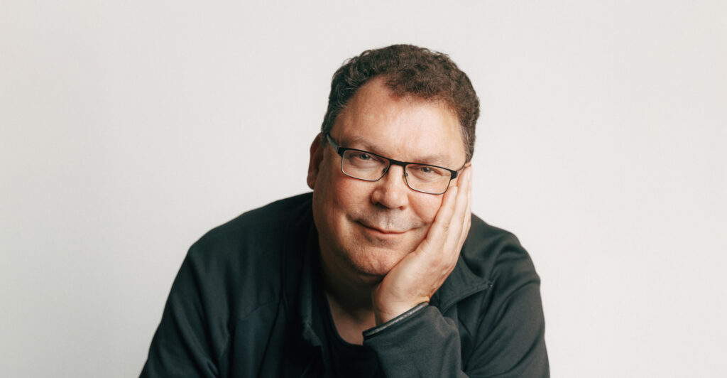 Eine Person mit Brille schaut freundlich in die Kamera. Sie stützt den Kopf mit einer Hand ab und trägt dunkle Kleidung. Der Hintergrund ist hell.