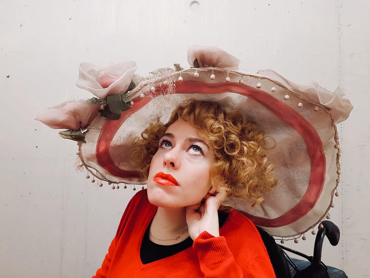 Eine Frau schaut nach oben. Sie trägt einen großen Hut und einen roten Pullover. Ihre Lippen sind rot geschminkt.