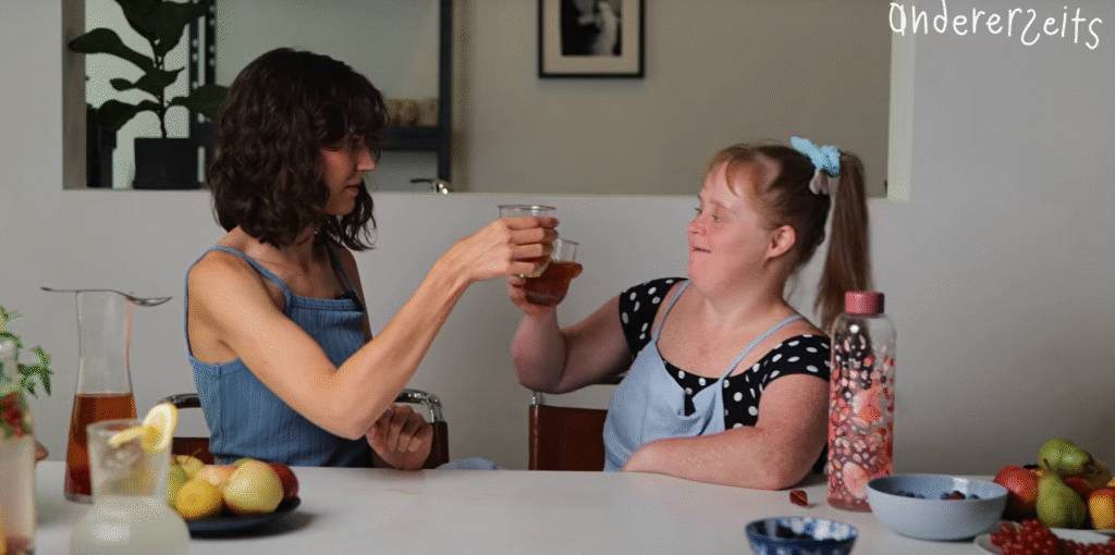 Mona und Hannah sitzen an einem Tisch und Prosten sich zu. Auf dem Tisch liegt Obst und Gefäße mit verschiedenen Getränken-