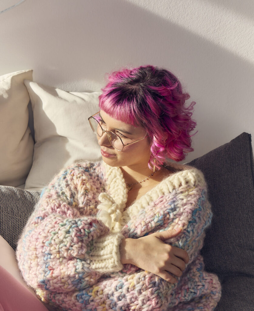 Eine Person mit kurzen pinken Haaren sitzt auf einem Sofa. Sie trägt eine bunte Strickjacke und trägt eine Brille.