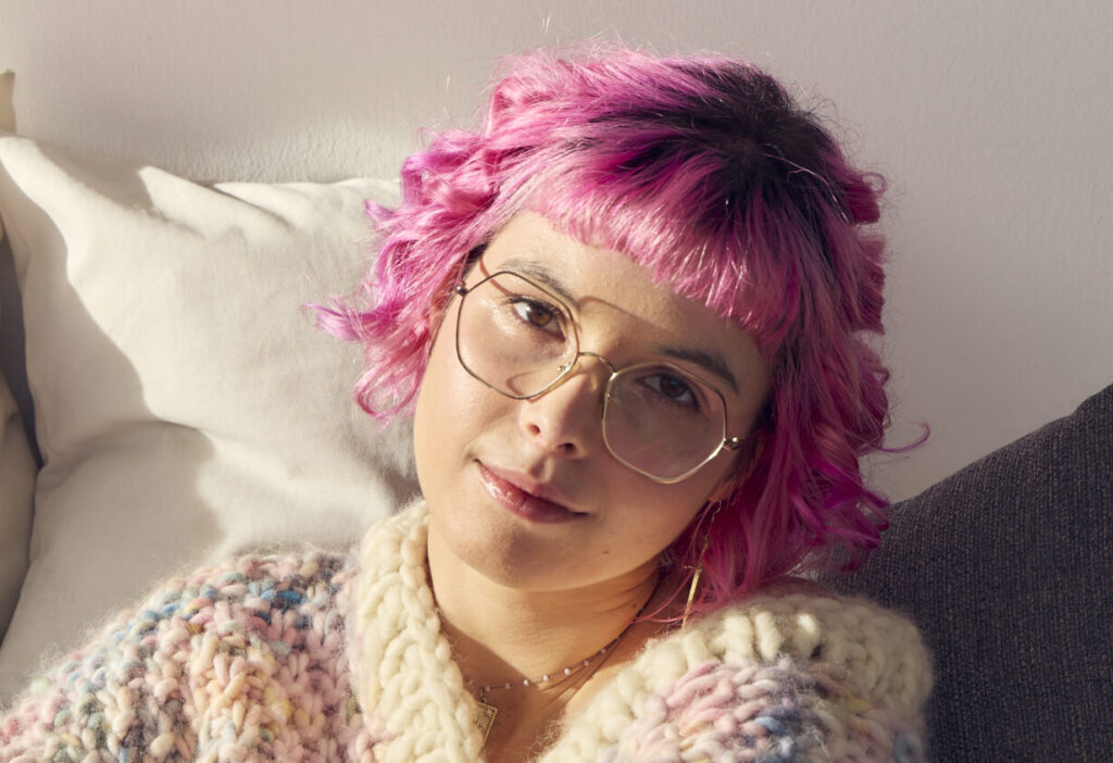 Eine junge Frau schaut mit freundlichen Blick in die Kamera. Sie trägt eine Brille und und hat kurze pinke Haare.