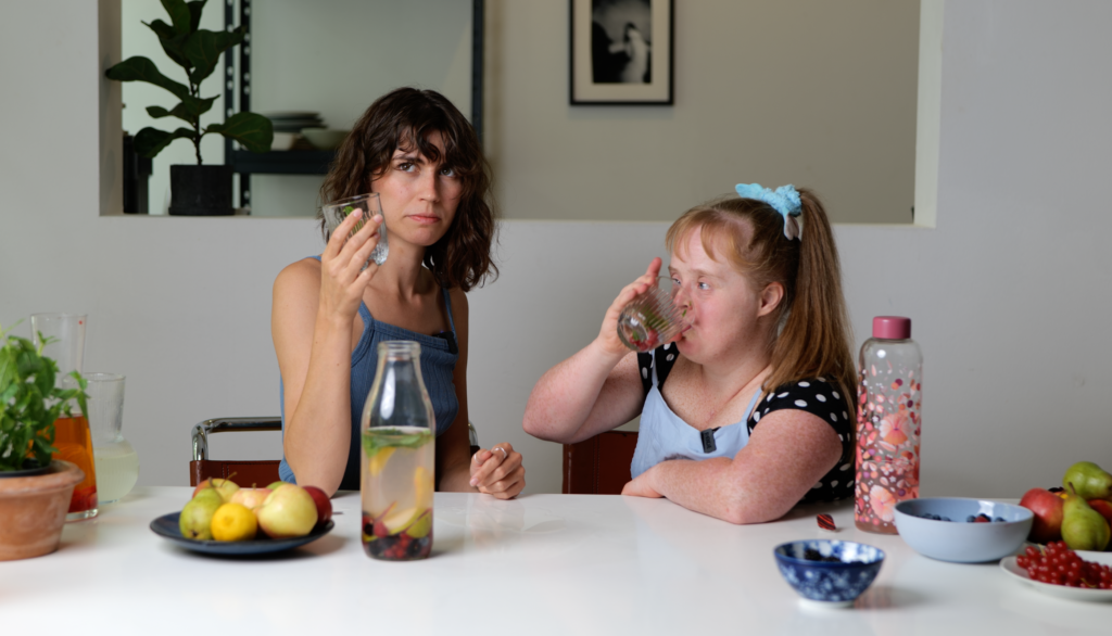Das Bild zeigt zwei Frauen, die auf einem großen Tisch sitzen. Beide halten ein Glas Wasser in der Hand. Auf dem Tisch stehen viele Früchte und weitere Flaschen mit Wasser.