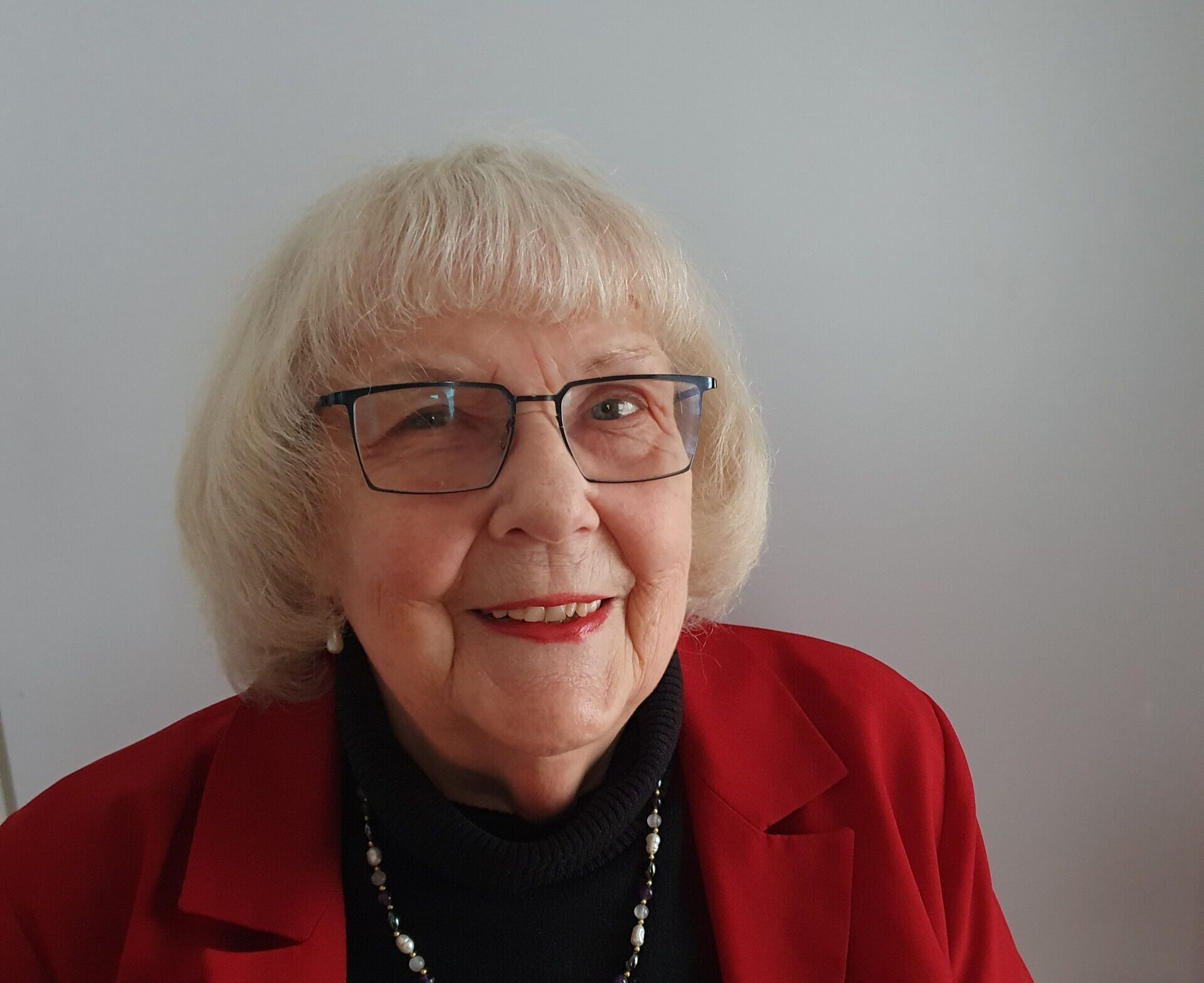 Eine Frau mit freundlichen Blick in die Kamera. Sie trägt eine Brille und hat kurze weiße Haare. Sie trägt einen roten Blazer.