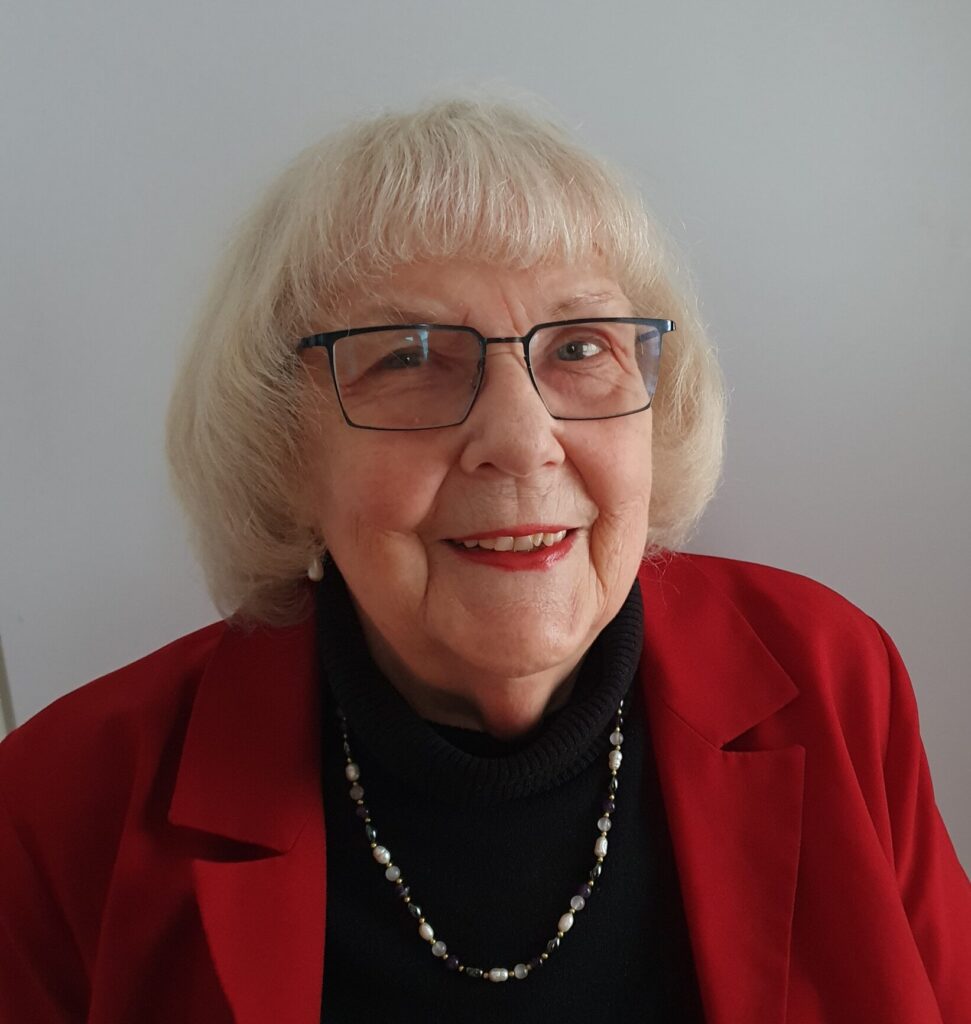 Eine Frau mit freundlichen Blick in die Kamera. Sie trägt eine Brille und hat kurze weiße Haare. Sie trägt einen roten Blazer.