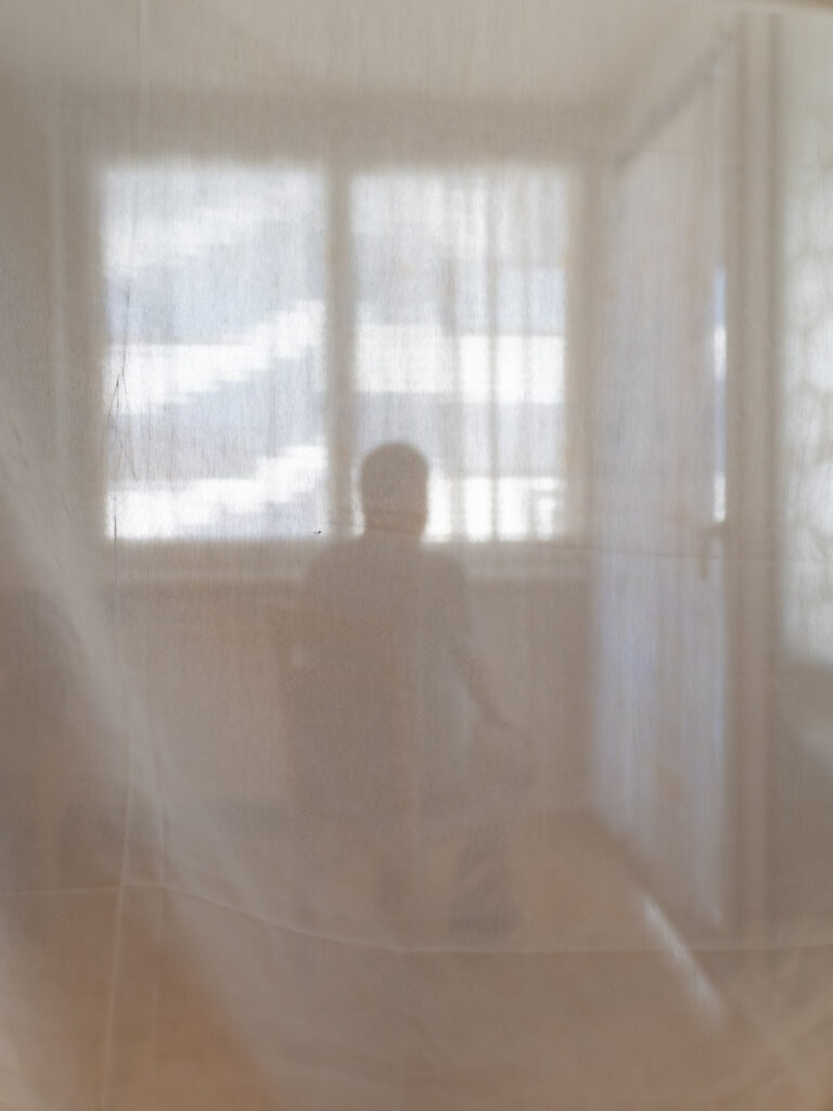 Eine Person sitzt hinter einem Stoffvorhang. Sie schaut aus dem Fenster.