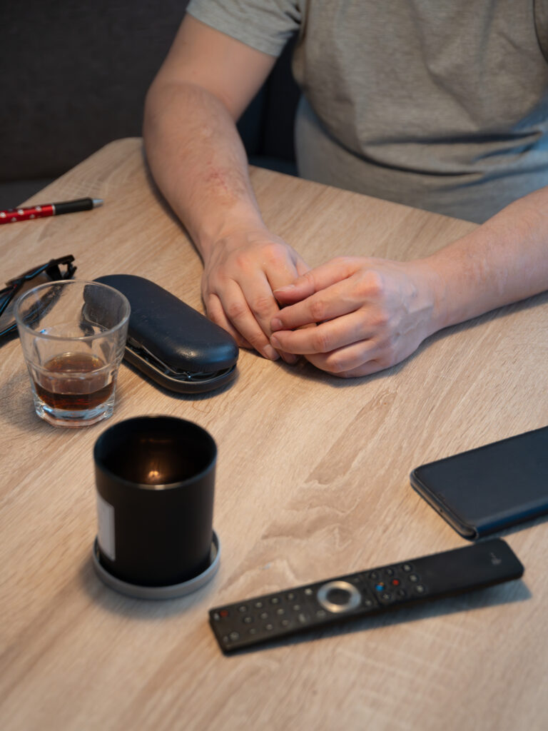 Eine Person sitzt an einem Tisch. Auf dem Tisch stehen mehrere Gegenstände, wie eine Fernbedienung, ein Glas Cola und eine Kerze.