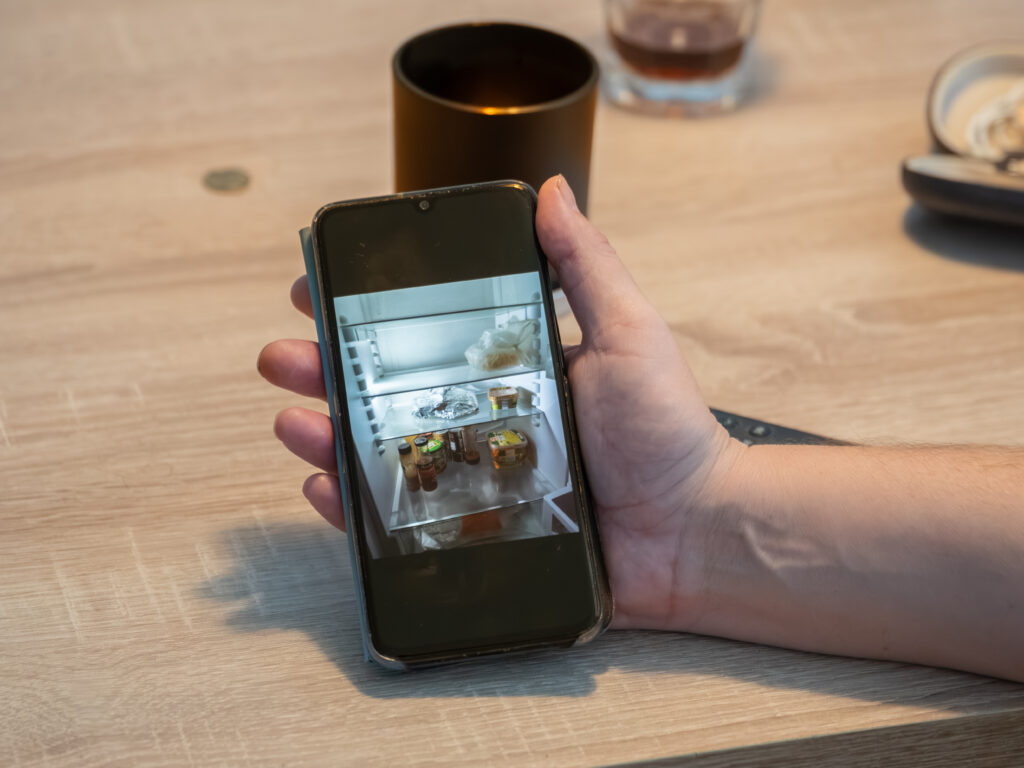Ein Mann hält ein Smartphone in der Hand. Darauf sieht man einen offenen Kühlschrank, der karg befüllt ist.