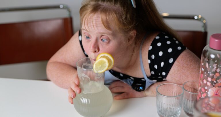 Eine junge Frau beugt sich über eine Karaffe mit Wasser. Auf der Karaffe sind zwei Zitronen-Scheiben. Auf dem Tisch stehen zwei Gläser und eine Wasserflasche.