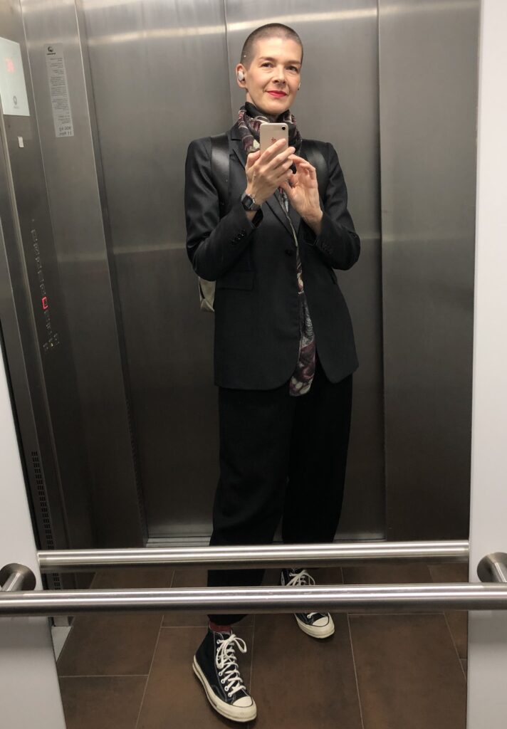 Eine Frau macht eine Selfie von sich in einem Spiegel. Sie trägt einen Blazer und hat kurze Haare. Sie trägt Lippenstift und Kopfhörer.