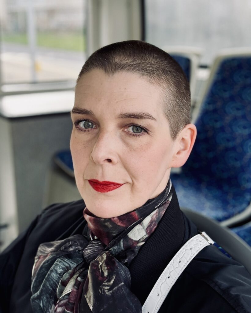 Eine Frau schaut mit ernstem Blick in die Kamera. Sie hat kurze, dunkle Haare. Sie trägt roten Lippenstift und ein Halstuch. Sie sitzt in einem Bus.