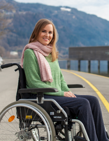 Eine Frau schaut mit freundlichen Blick in die Kamera. Sie trägt einen grünen Pullover und einen pinken Schal. Sie nützt einen Rollstuhl. Im Hintergrund sin Berge und ein See.