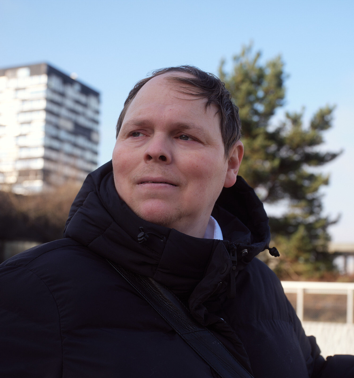 Ein Manns schaut mit ernstem Blick nach links. Er hat kurze braune Haare und trägt eine schwarze Jacke.