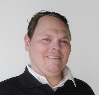 Ein Mann schaut mit freundlichen Blick in die Kamera. Er hat kurze braune Haare und trägt ein Hemd.