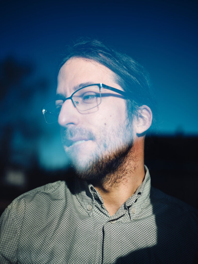 Ein Mann schaut mit ernsten Blick nach links. Er steht hinter einer Fensterscheibe und trägt eine Brille.