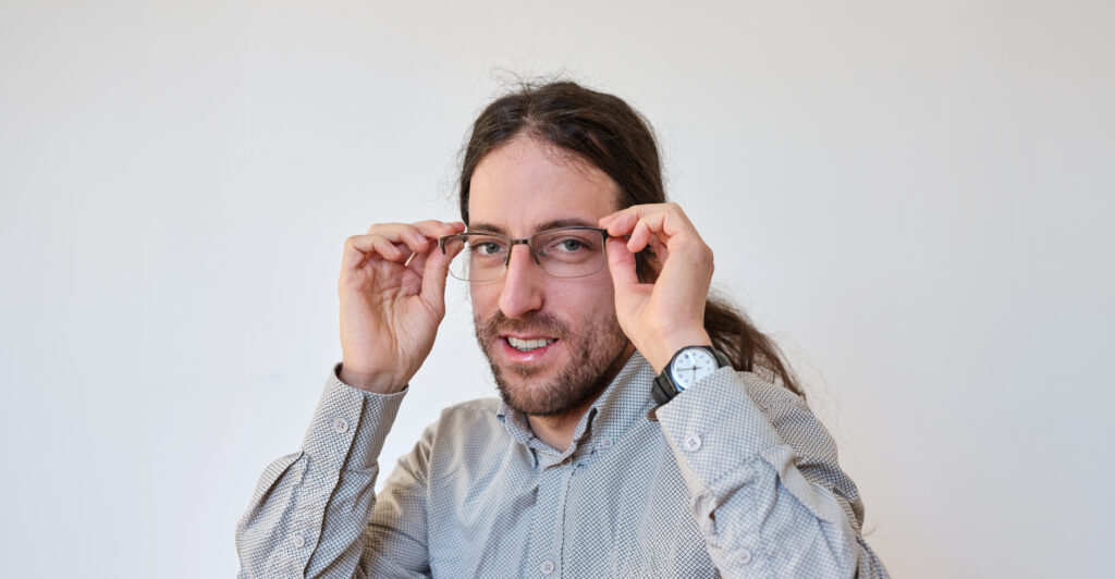 EIn Mann schaut mit freundlichen Blick in die Kamera. Er trägt eine Brille und hat die Haare zu einem Zopf gebunden.