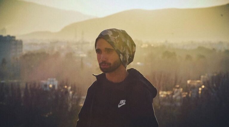 Eine Person schaut mit ernsten Blick nach links. Sie trägt eine Mütze und eine schwarze Jacke. Im Hitnergrund sind mehrere Häuser und Berge zu sehen.