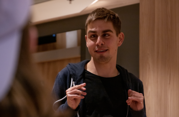 Ein junger Mann schaut nach links. Er hat kurze braune Haare und trägt einen blauen Pullover.