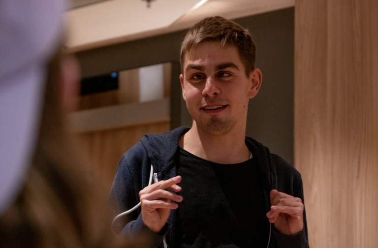 Ein junger Mann schaut nach links. Er hat kurze braune Haare und trägt einen blauen Pullover.