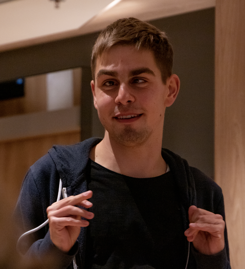 Ein junger Mann schaut nach links. Er hat kurze braune Haare und trägt einen blauen Pullover.