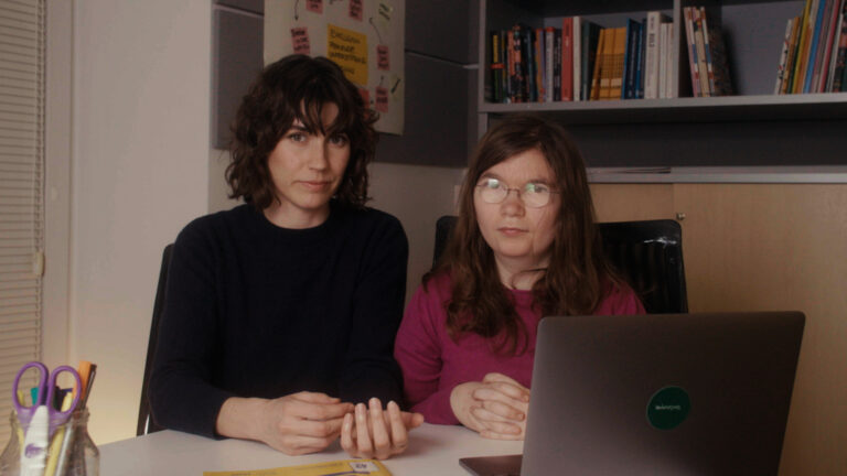 Zwei Frauen sitzen auf einen Tisch und schauen mit ernsten Blick in die Kamera. Vor ihnen steht ein offener Laptop. Im Hintergrund steht ein Bücherregal.