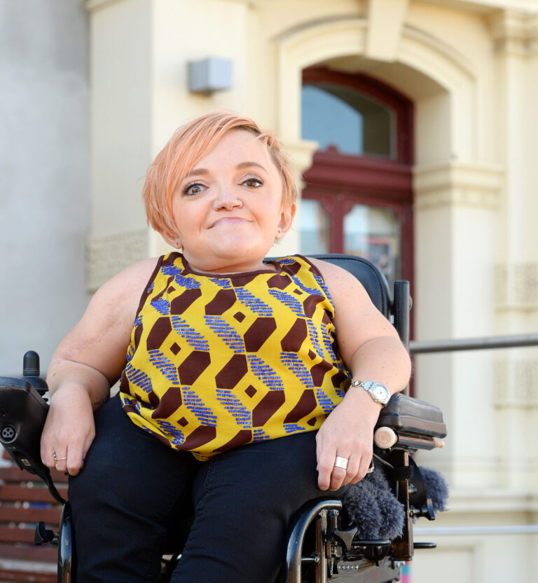 Eine Frau schaut m it freundlichen Blick in die Kamera. Sie hat kurze Haare und benutzt einen Rollstuhl.