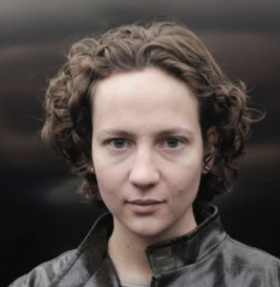 Eine Frau schaut mit ernsten Blick in die Kamera. Sie hat braune kurze Locken.