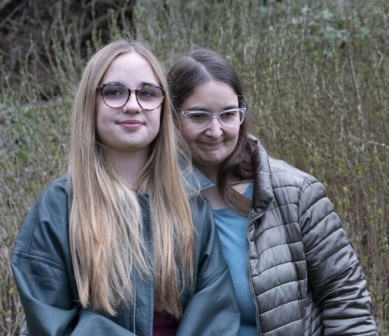 Zwei Frauen stehen nebeneinander. Links hat blonde lange Haare und trägt eine blaue Jacke. Die Frau rechts hat braune Haare, trägt eine Brille und eine braune Jacke.