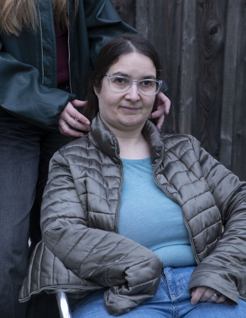 Eine Frau schaut mit ernsten Blick nach oben. Sie trägt eine braune Jacke. Im Hintergrund steht eine Person.