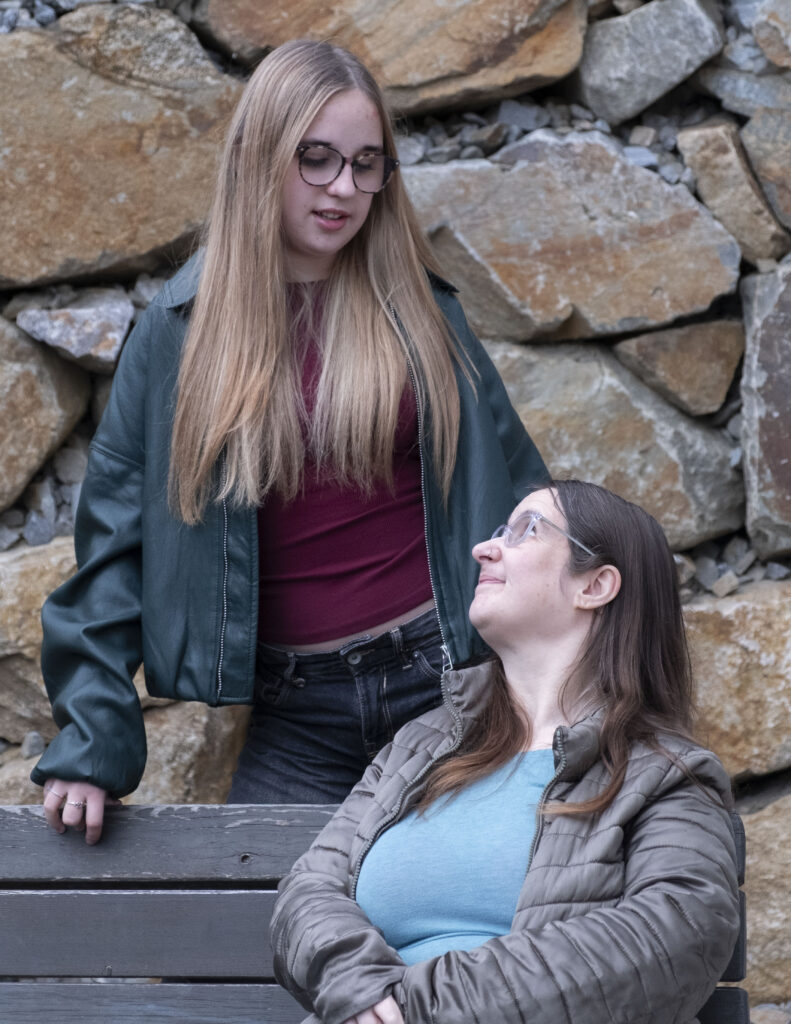 Zwei Frauen schauen sich gegenseitig an. Eine Frau sitzt rechts auf er Bank und schaut nach oben. Die andere Frau steht hinter der Bank und schaut nach unten.