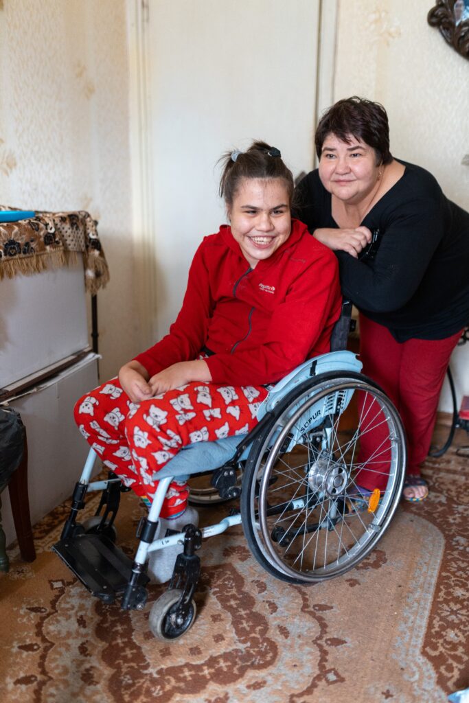 Ein Mädchen sitzt in einem Rollstuhl und schaut mit freundlichen Blick nach rechts. Dahinter steht eine Frau, sie lehnt sich über den Rollstuhl.