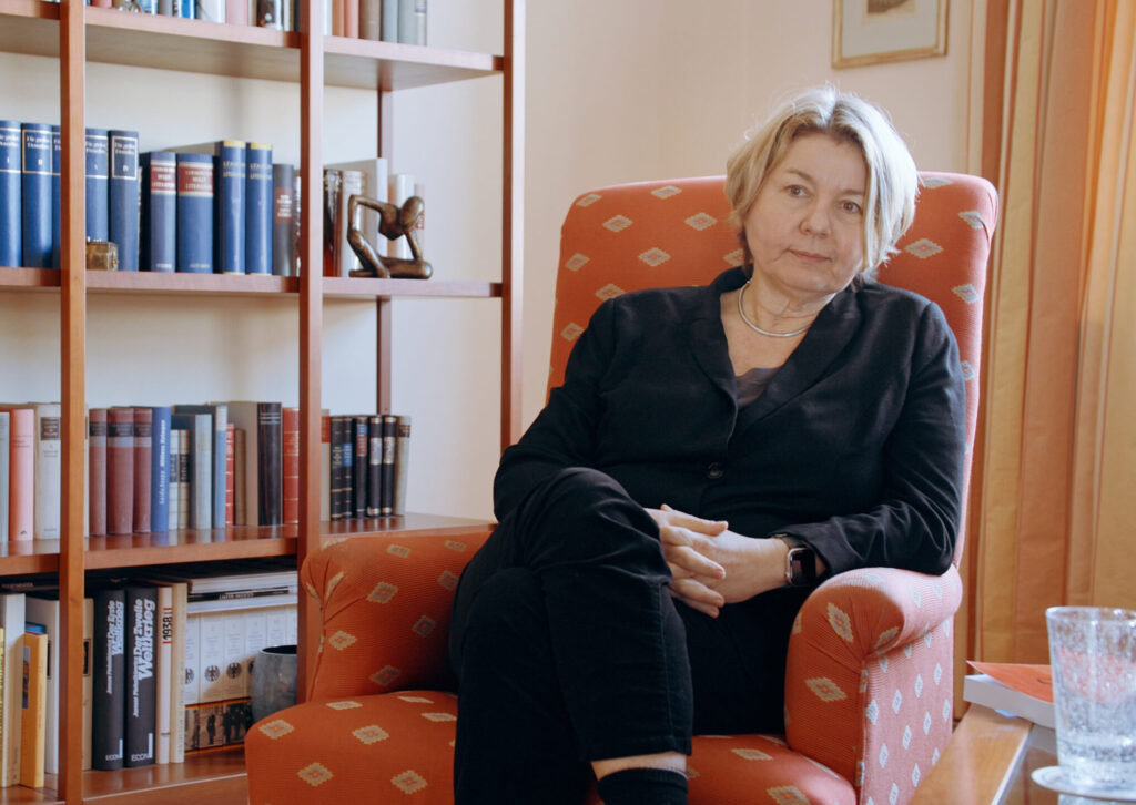 Eine Frau mit kurzen blonden Haaren schaut mit ernsten Blick in die Kamera. Sie sitzt auf einem roten Stuhl und trägt schwarze Kleidung. Im Hintergrund steht ein Bücherregal.