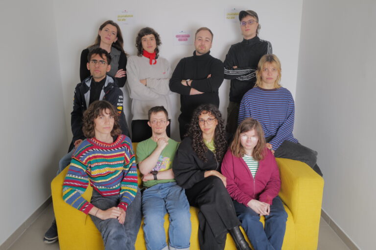 Das Team von andererseits sitzt und steht rund um ein gelbes Sofa. Alle blicken ernst in die Kamera. Es sind Menschen mit und ohne Behinderungen, Männer und Frauen.