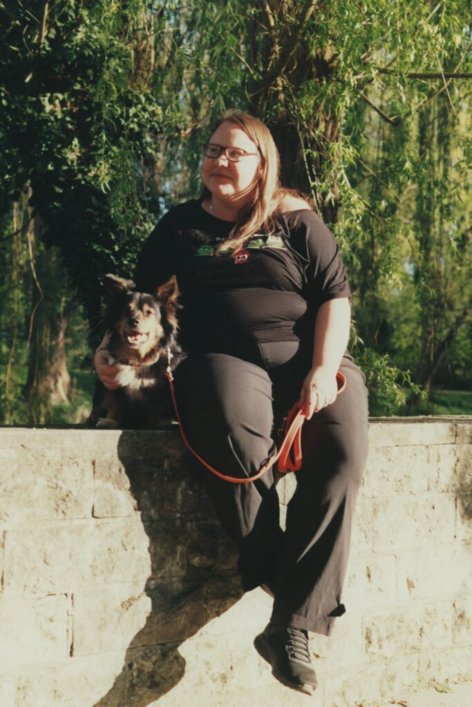 Eine Frau sitzt auf einer Mauer. Links von ihr sitzt ein Hund. Sie trägt schwarze Kleidung. Sie hat dunkelblonde lange Haare und trägt eine Brille.