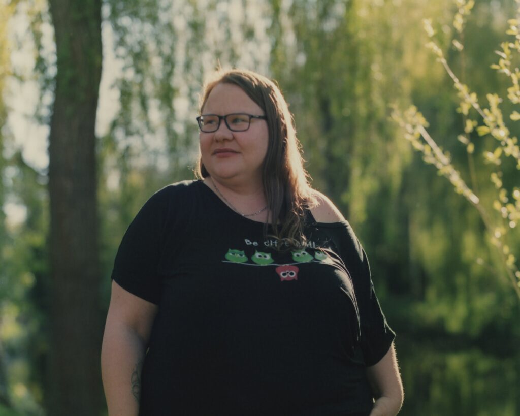 Eine Frau steht in einem Park. Sie hat dunkelblonde lange Haare und trägt eine Brille. Sie trägt schwarze Kleidung und schaut mit dem Kopf nach links.