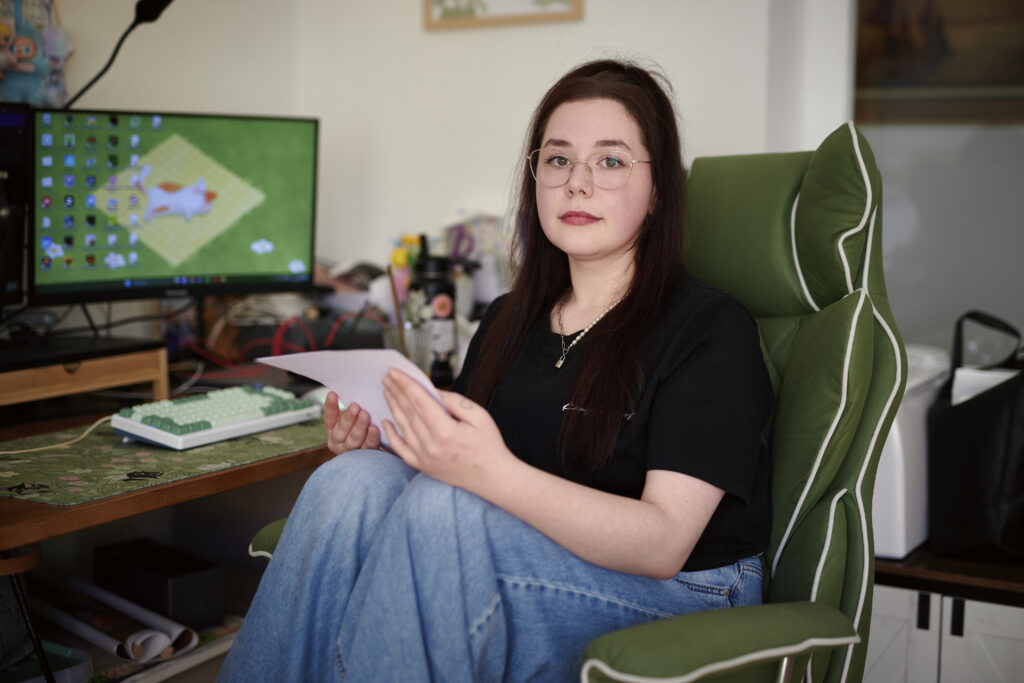 Eine junge Frau schaut mit ernstem Blick in die Kamera. Sie sitzt in einem grünen Bürostuhl und hält ein Blatt Papier in den Händen. Im Hintergrund sieht man einen Computer-Bildschirm.